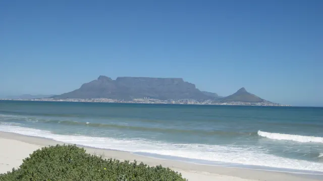 White surf reaches pale sand below a distant flat mountain.