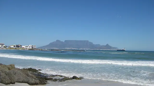 A distant flat-topped mountain appears beyond white water and shoreline rocks.