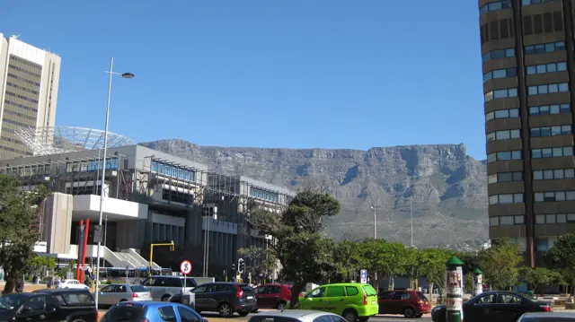 City traffic passes a modern building below a flat mountain ridge.