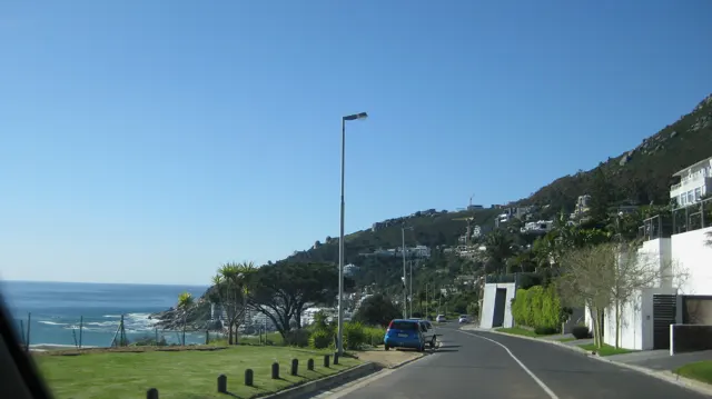 A road follows hillside homes above the blue sea.