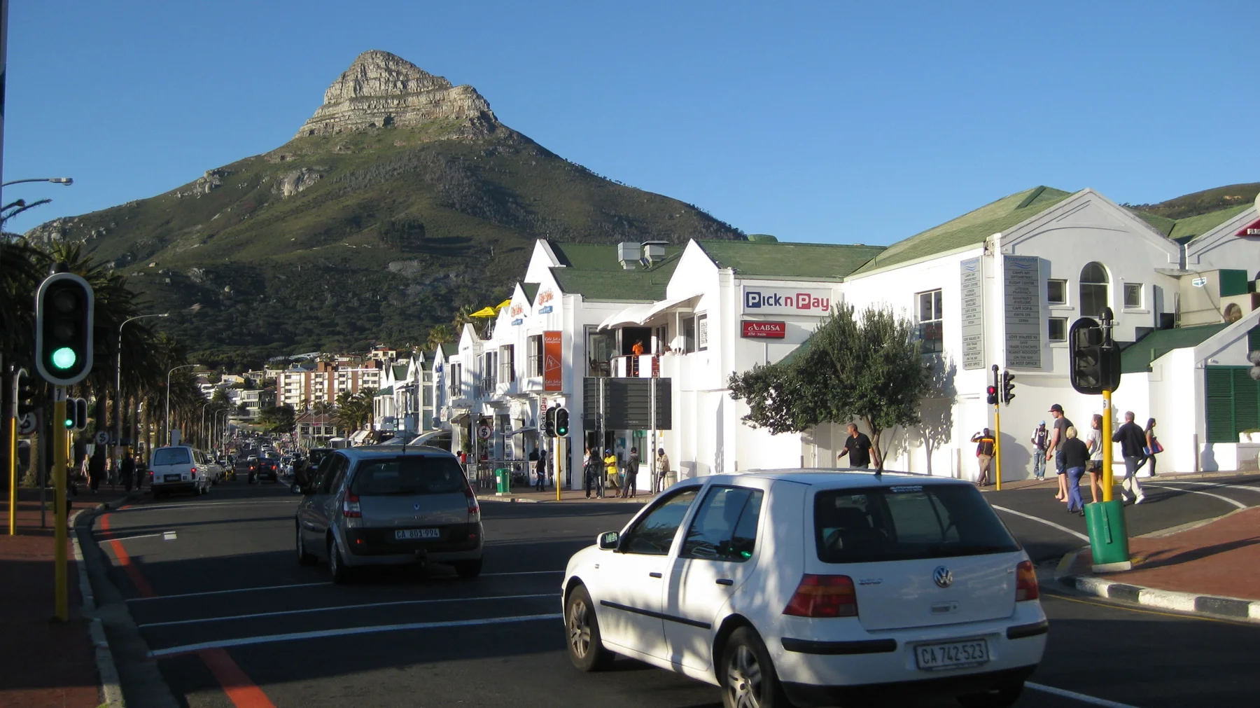 Cars pass white shops beneath a pointed mountain summit.