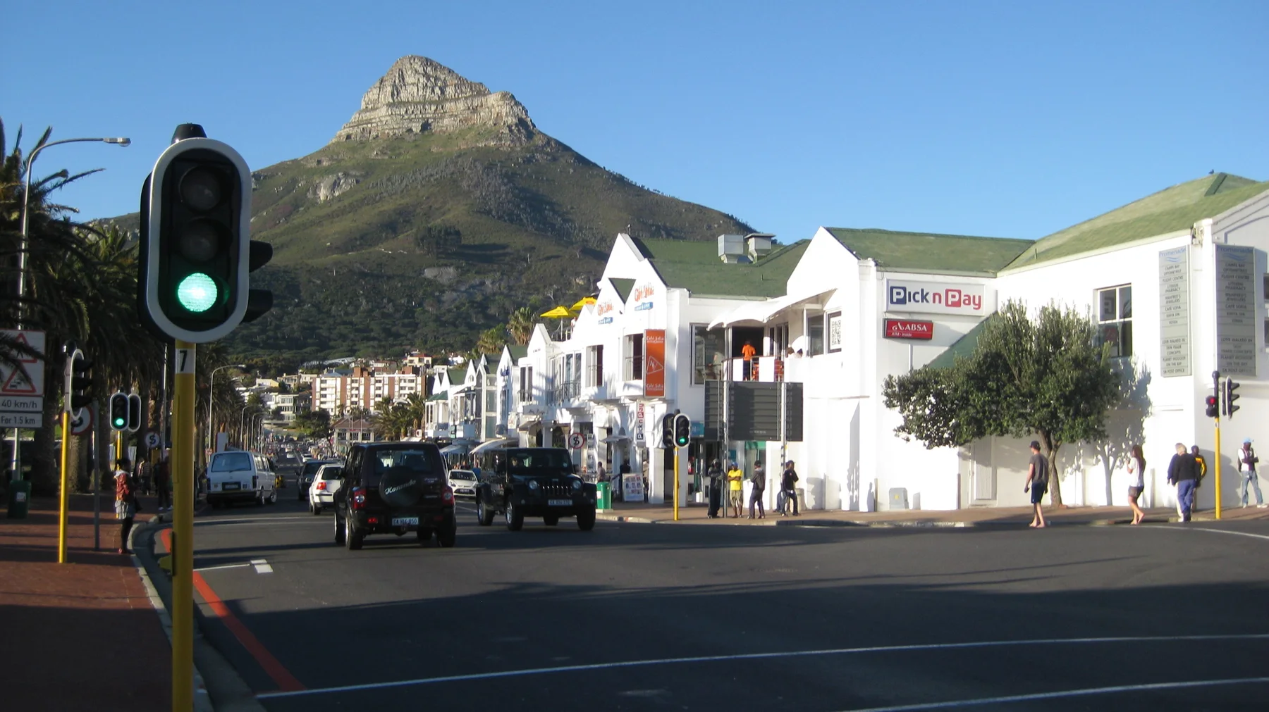 A green traffic light stands before white shops and a pointed peak.