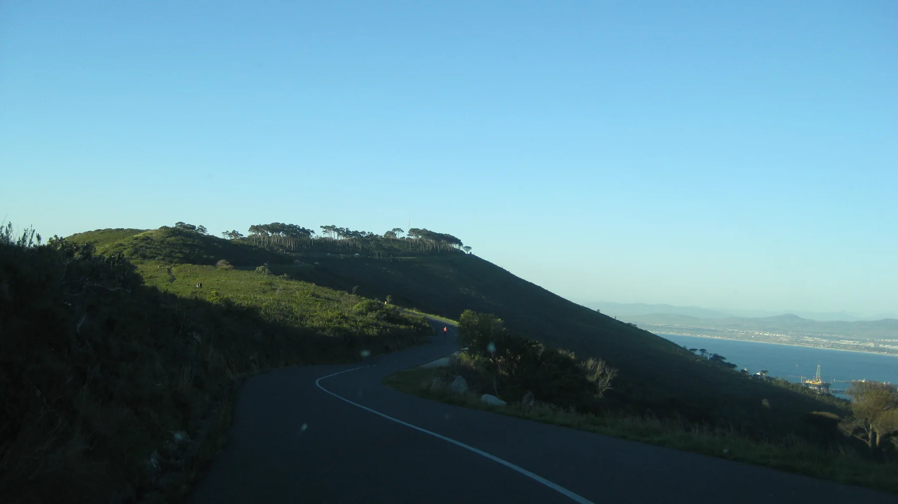 A road curves along a dark green hillside above distant water.
