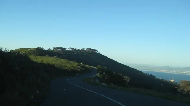 A road curves along a dark green hillside above distant water.