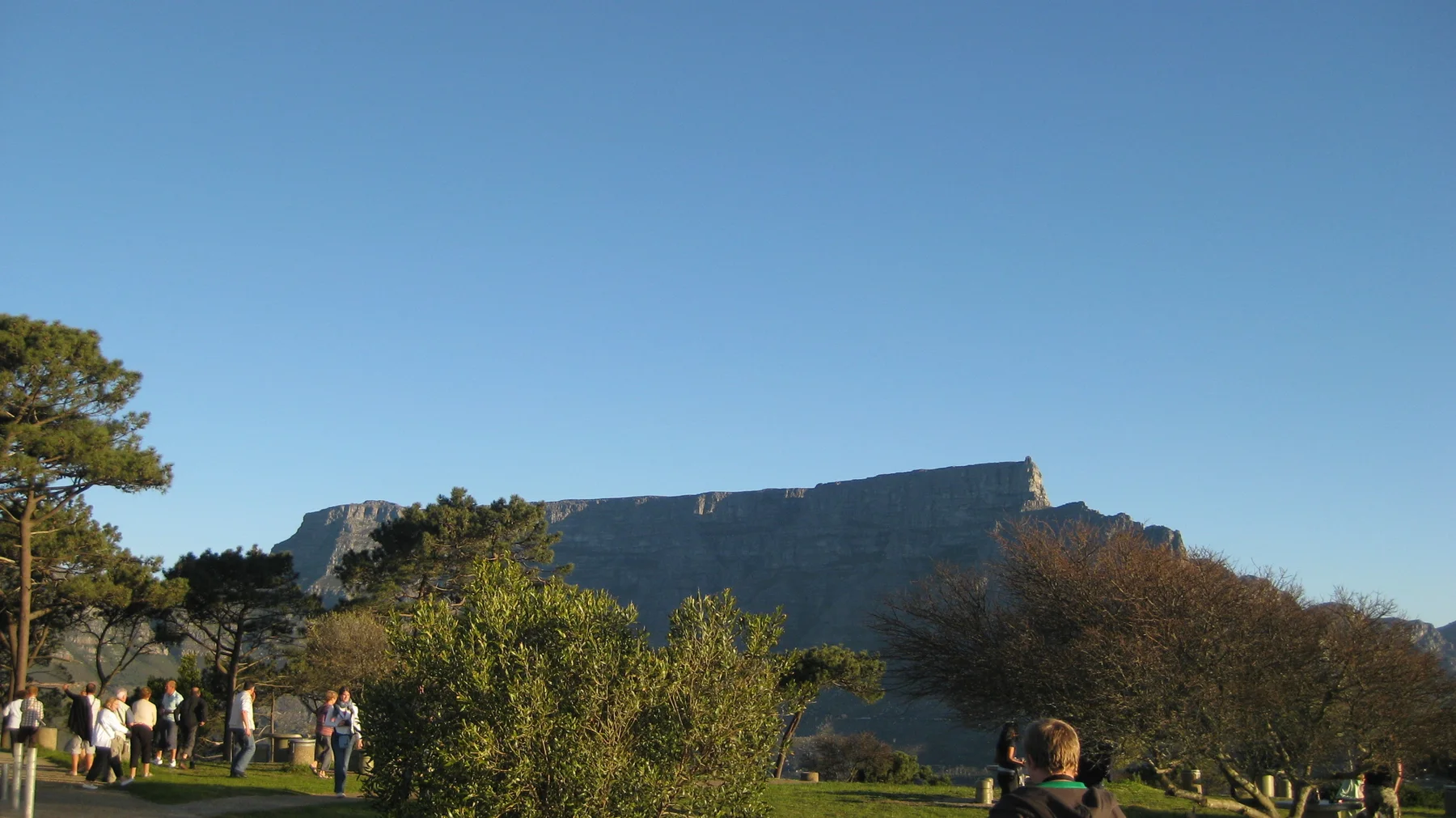 A flat mountain ridge rises beyond park trees and visitors.