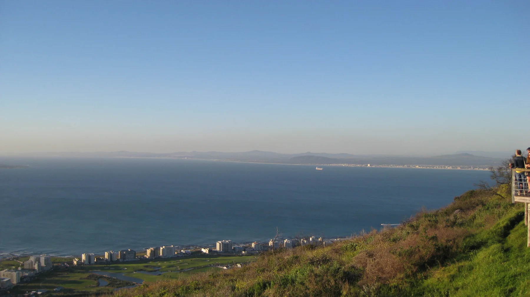 A grassy slope overlooks distant coastline under evening haze.