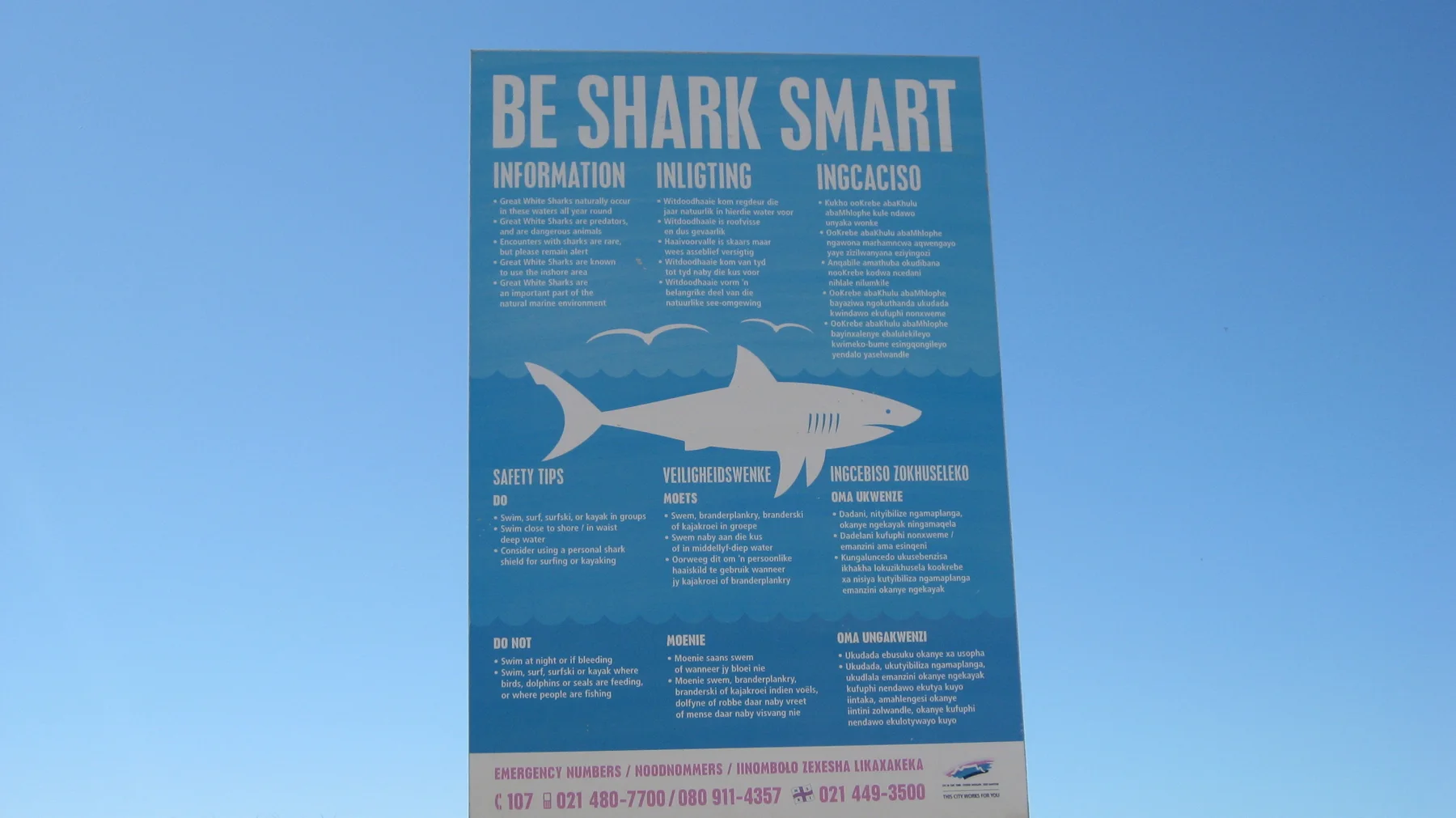 A blue beach information board is headed Be Shark Smart.