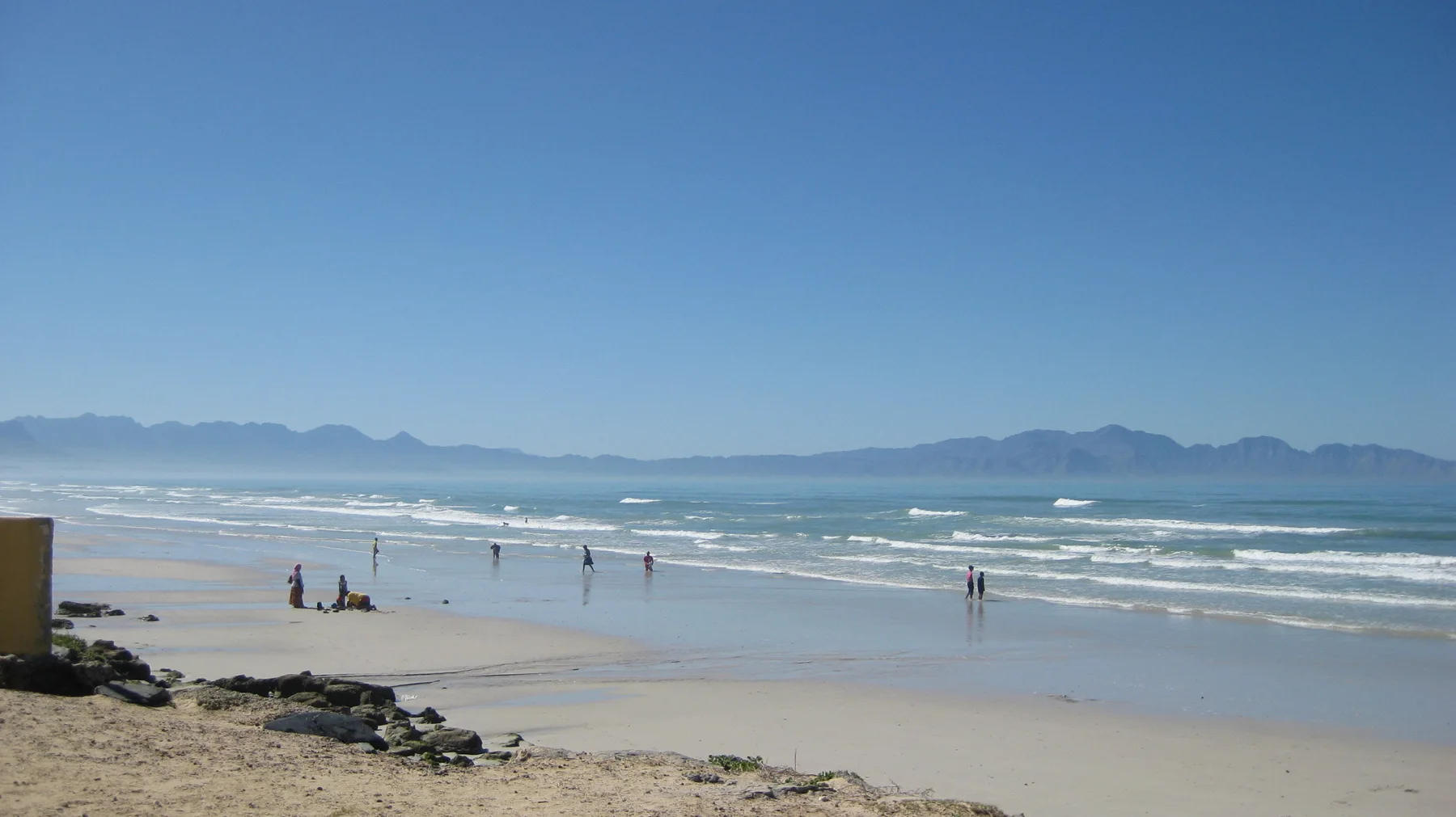 People walk and stand along a shallow sandy shoreline.