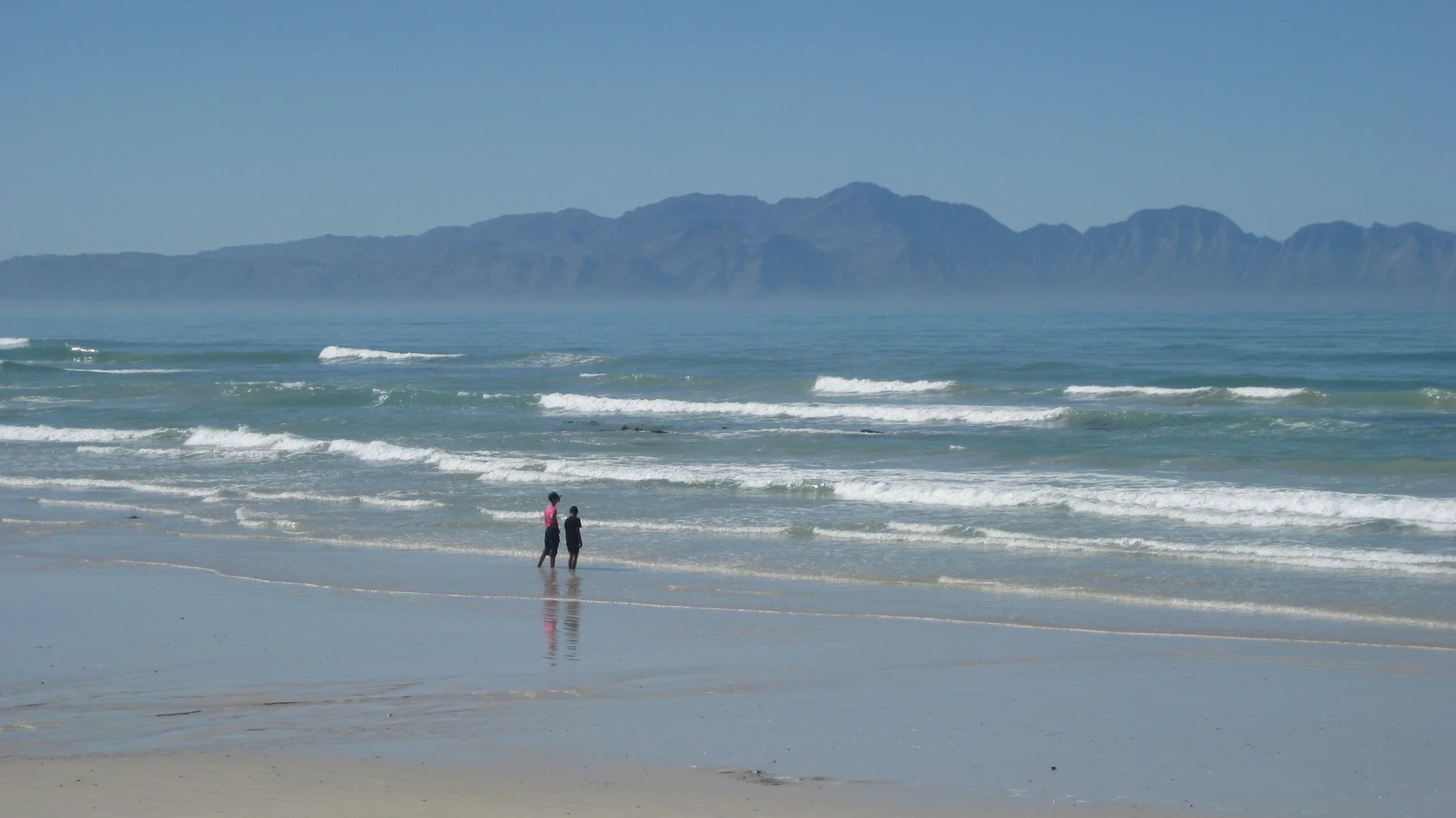 Two people stand in shallow surf below distant mountains.