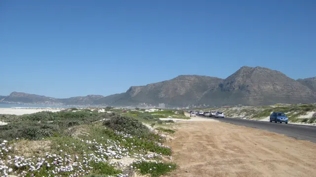 A sandy verge follows a coastal road beneath rugged mountains.
