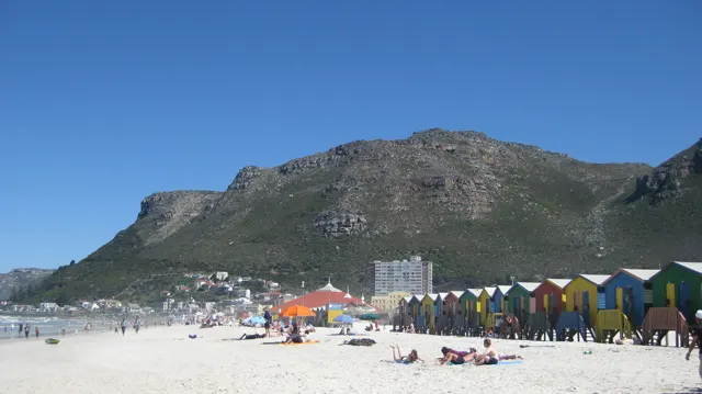 Colorful huts line a sunny beach beneath a rocky mountain.