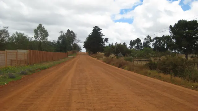 A red dirt road runs beside a long fence and scattered trees.