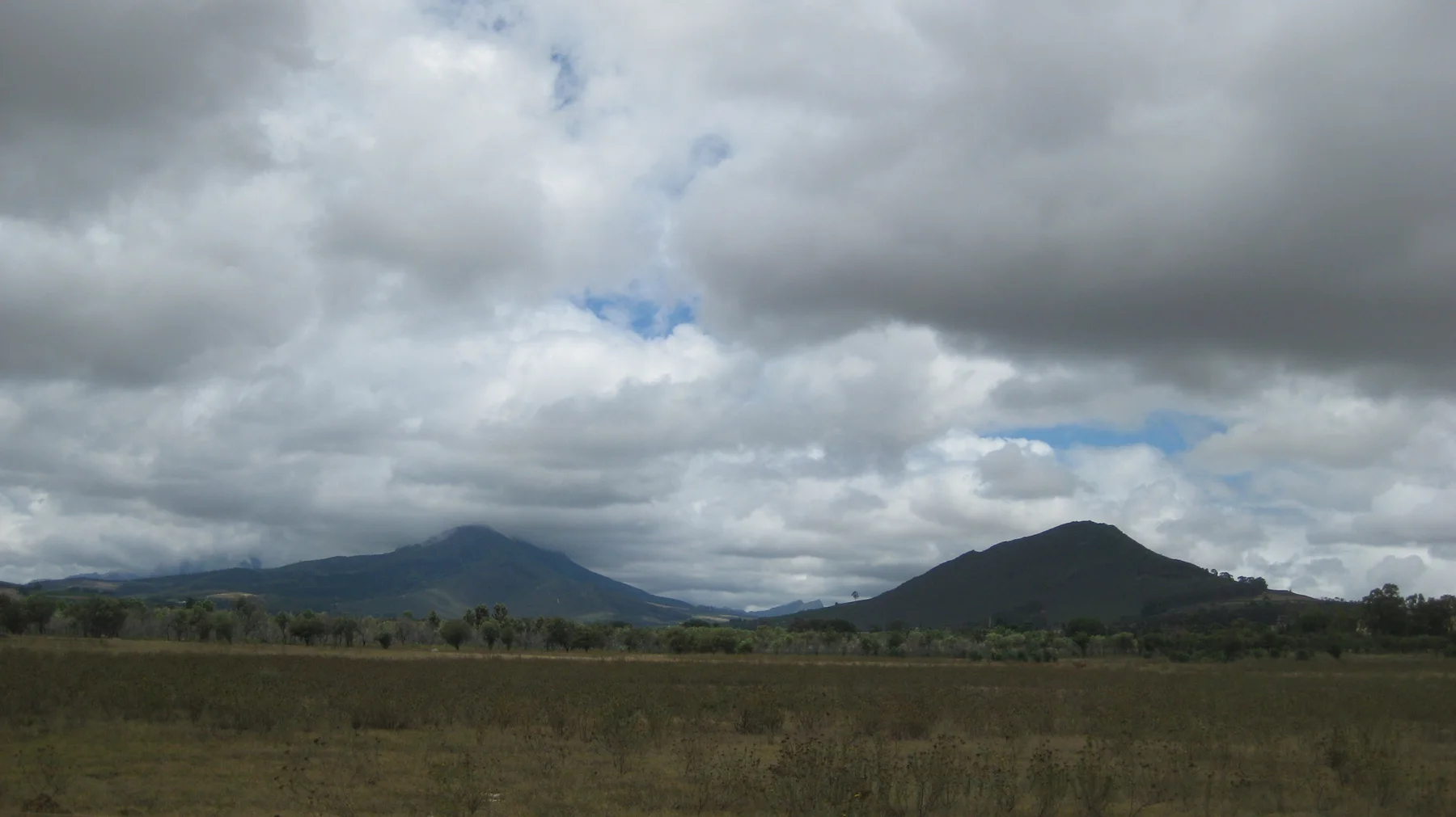 Two hills rise beyond open fields under heavy clouds.