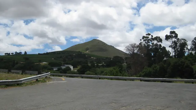 A broad road bend overlooks houses beneath a green hill.