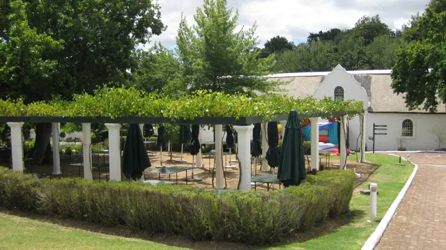 A vine-covered pergola shelters tables beside a white building.