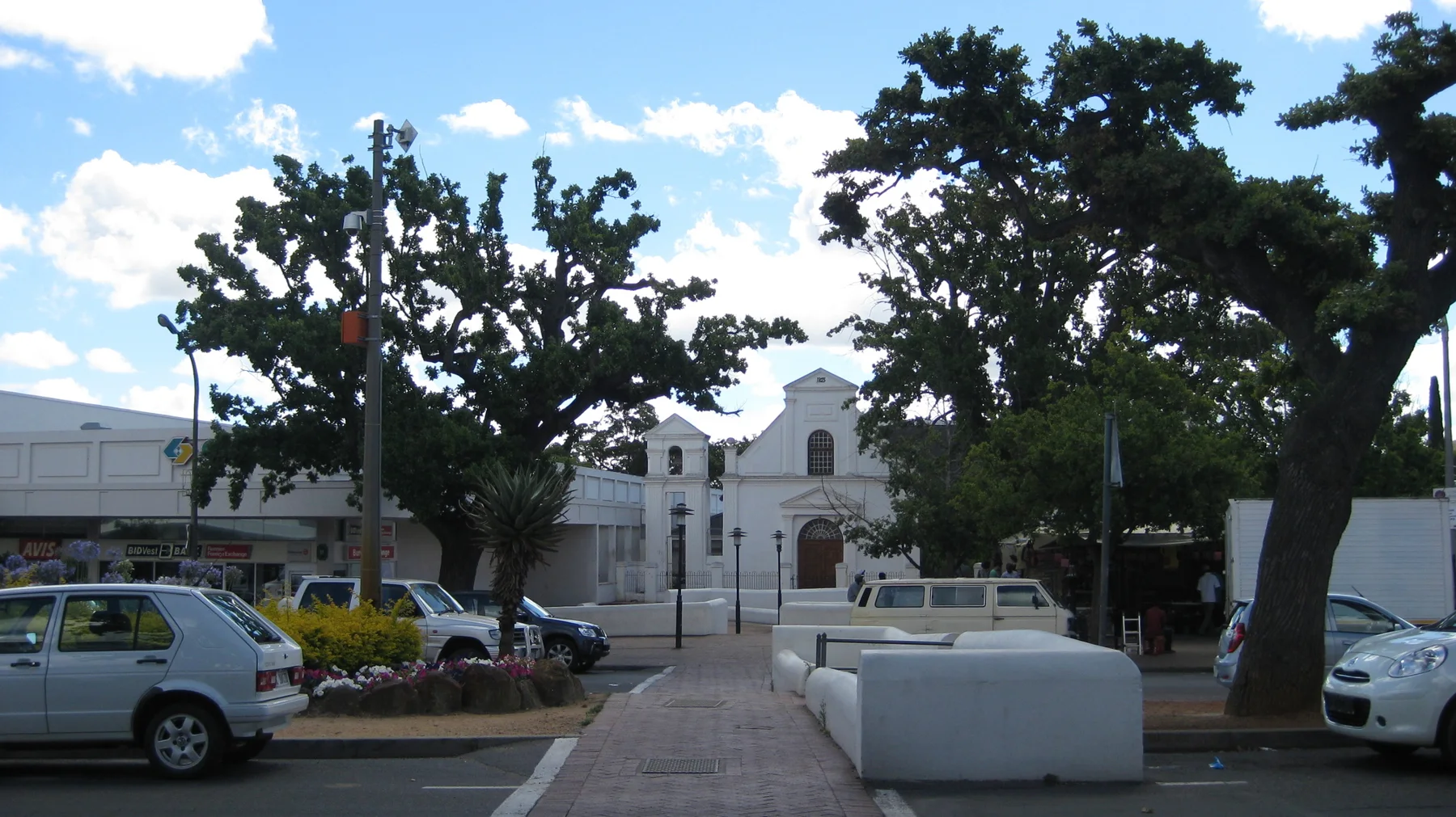 A white gabled building faces an open area with trees and parked cars.