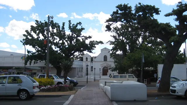 A white gabled building faces an open area with trees and parked cars.