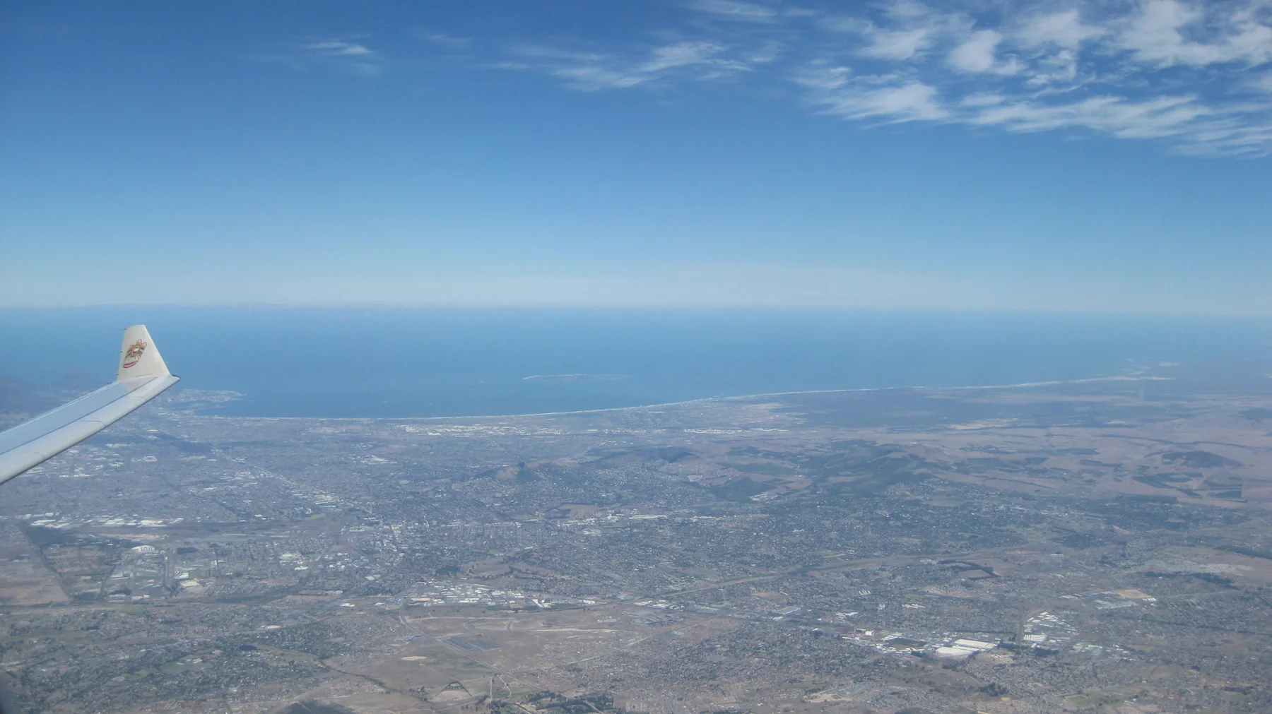 An aircraft wing frames a high view of city and blue coastline.