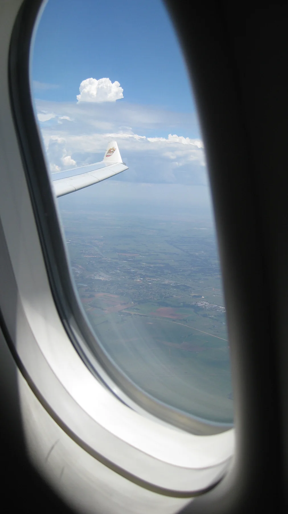An aircraft window frames blue sky, white clouds and a wing edge.