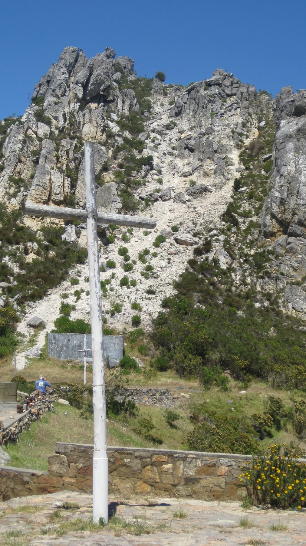 A white cross stands beneath a steep rocky slope.