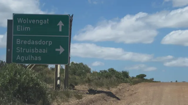A green direction sign stands above a broad dirt-road junction.