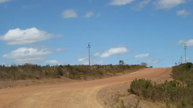 A red dirt track rises past scrub and utility poles.