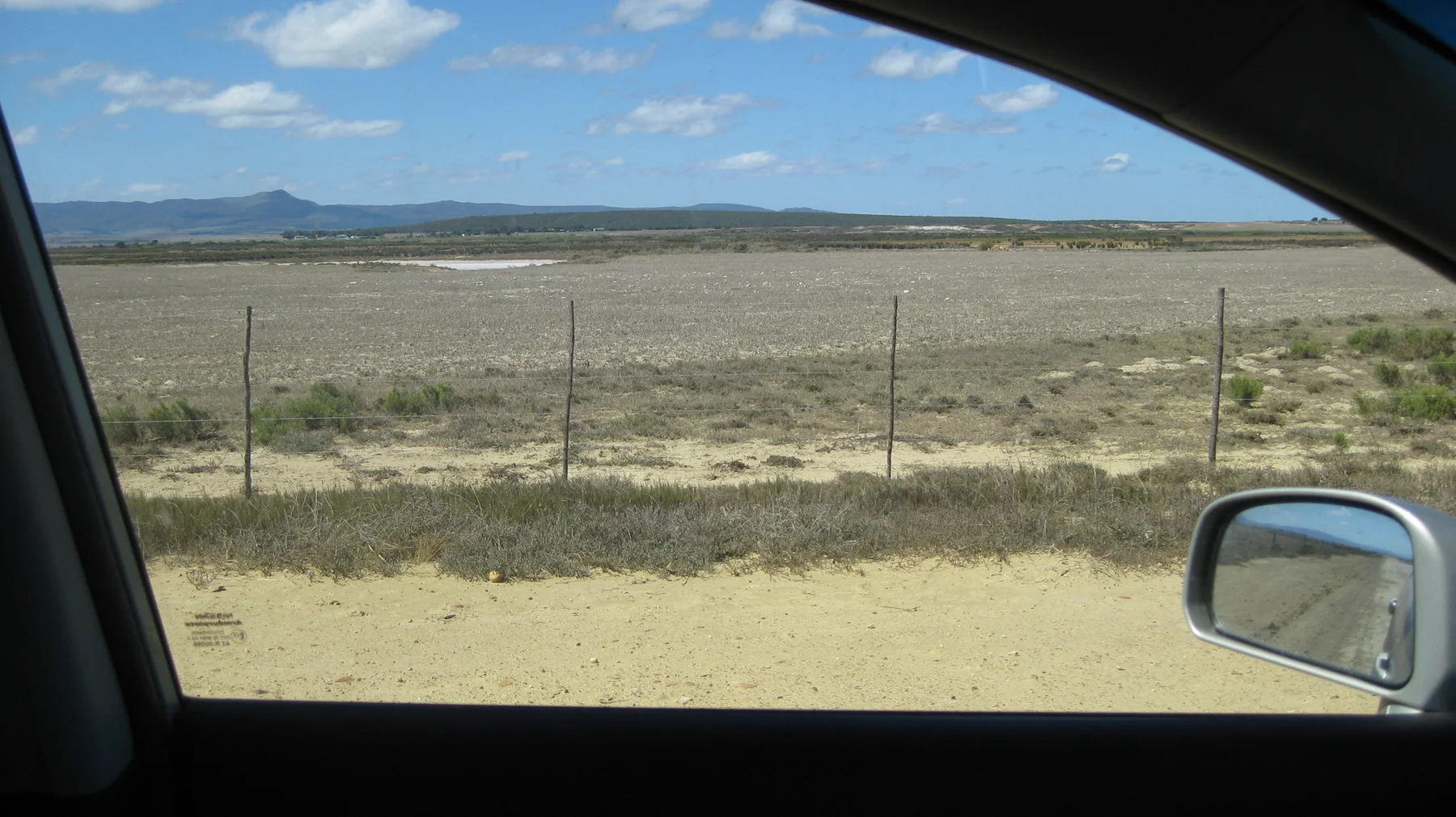 A car window frames a dry fenced field and distant hills.