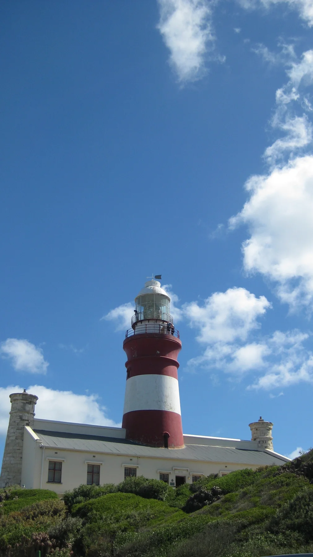 A red-and-white lighthouse rises above pale buildings and green ground.