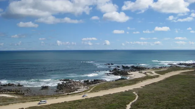 A pale road follows a rocky shore beside blue ocean.