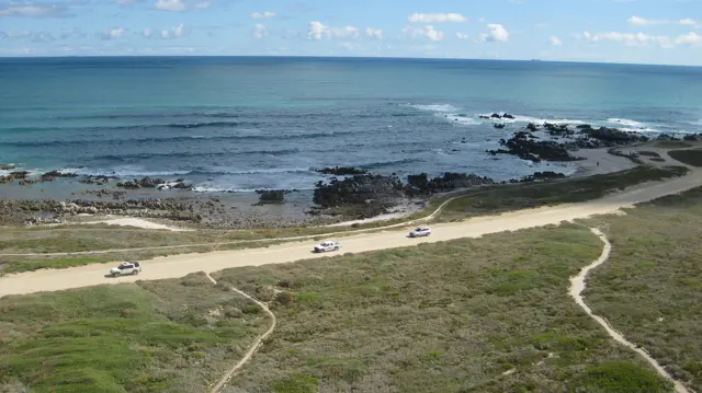 Narrow paths cross grass toward a road beside dark coastal rocks.