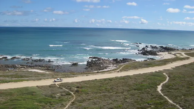 A shallow rocky bay lies beside a coastal track under white clouds.