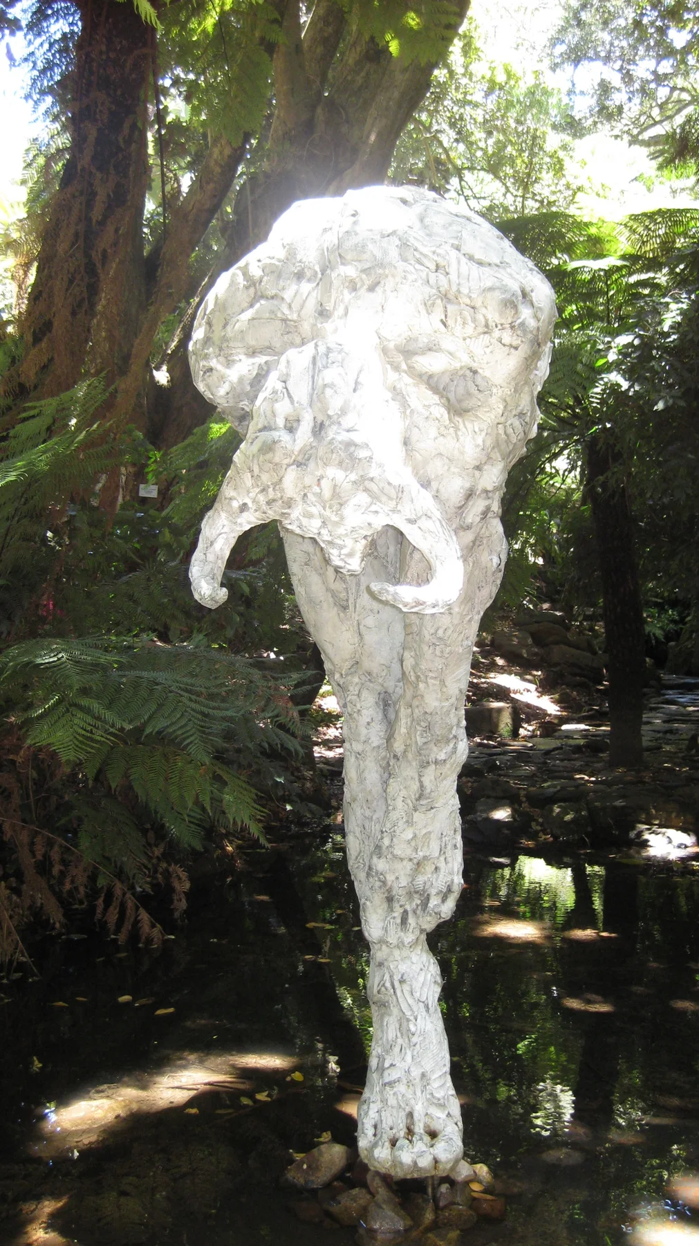 A tall pale abstract sculpture stands among shaded garden trees.