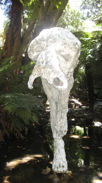 A tall pale abstract sculpture stands among shaded garden trees.