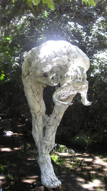 A pale sculptural form curves above a narrow base in garden shade.