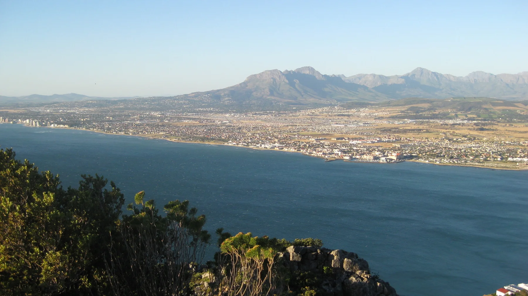 Foreground shrubs frame a developed coastal plain and blue bay.
