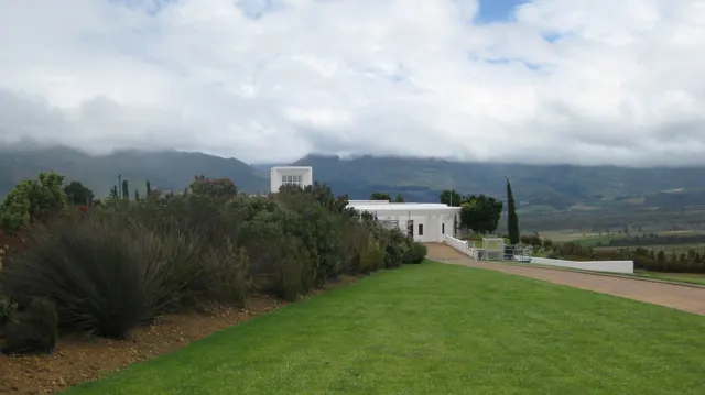A white building stands beyond a lawn under clouded mountains.