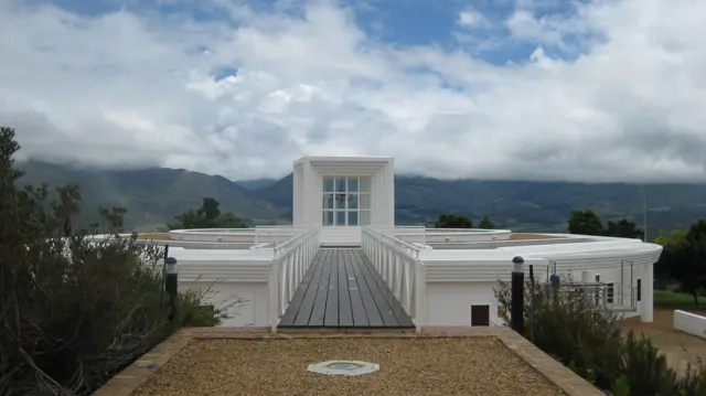 A geometric white building stands beneath cloud-covered mountains.