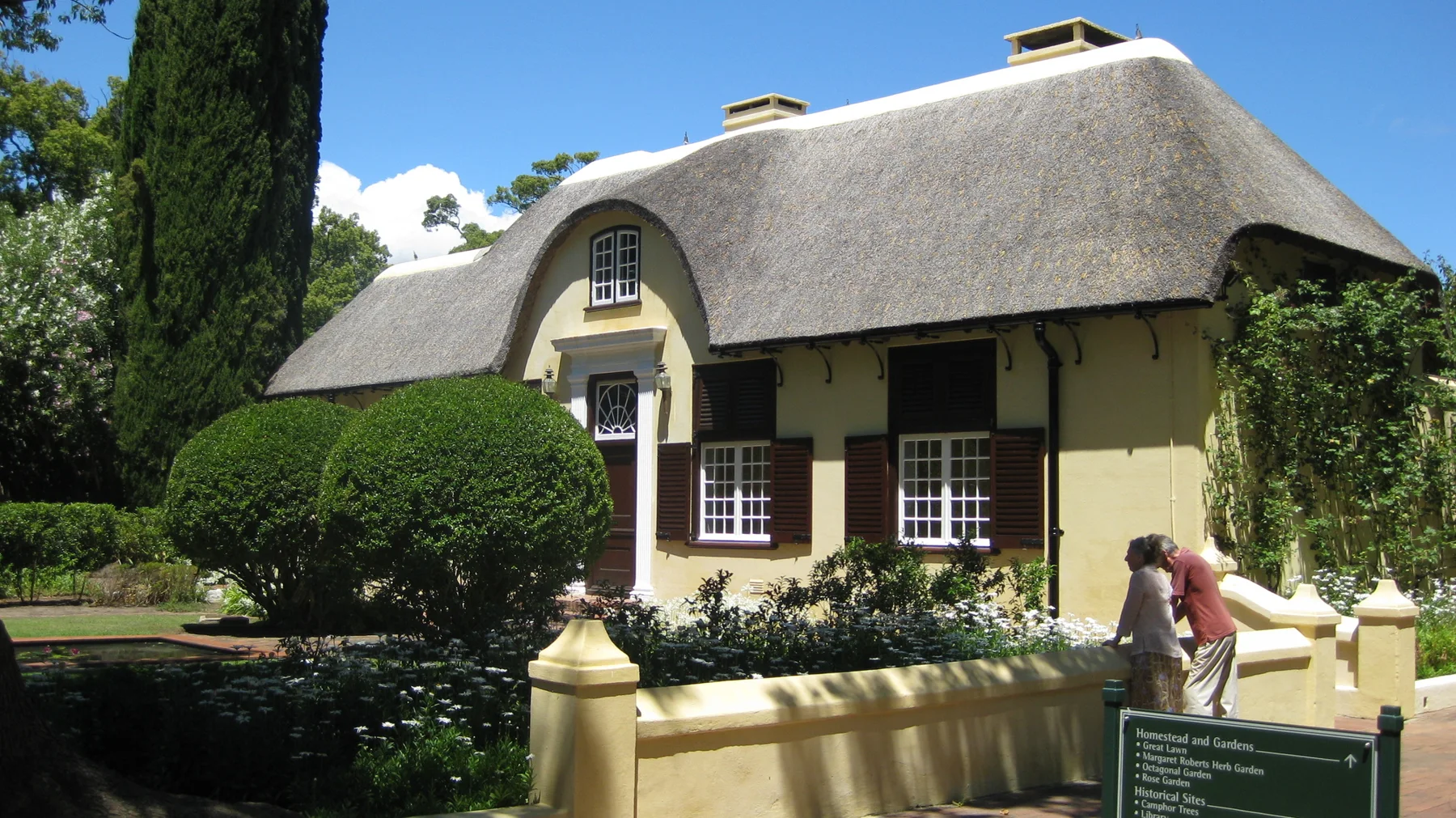 A pale yellow house with a curved thatched roof stands behind clipped shrubs.