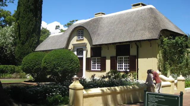 A pale yellow house with a curved thatched roof stands behind clipped shrubs.