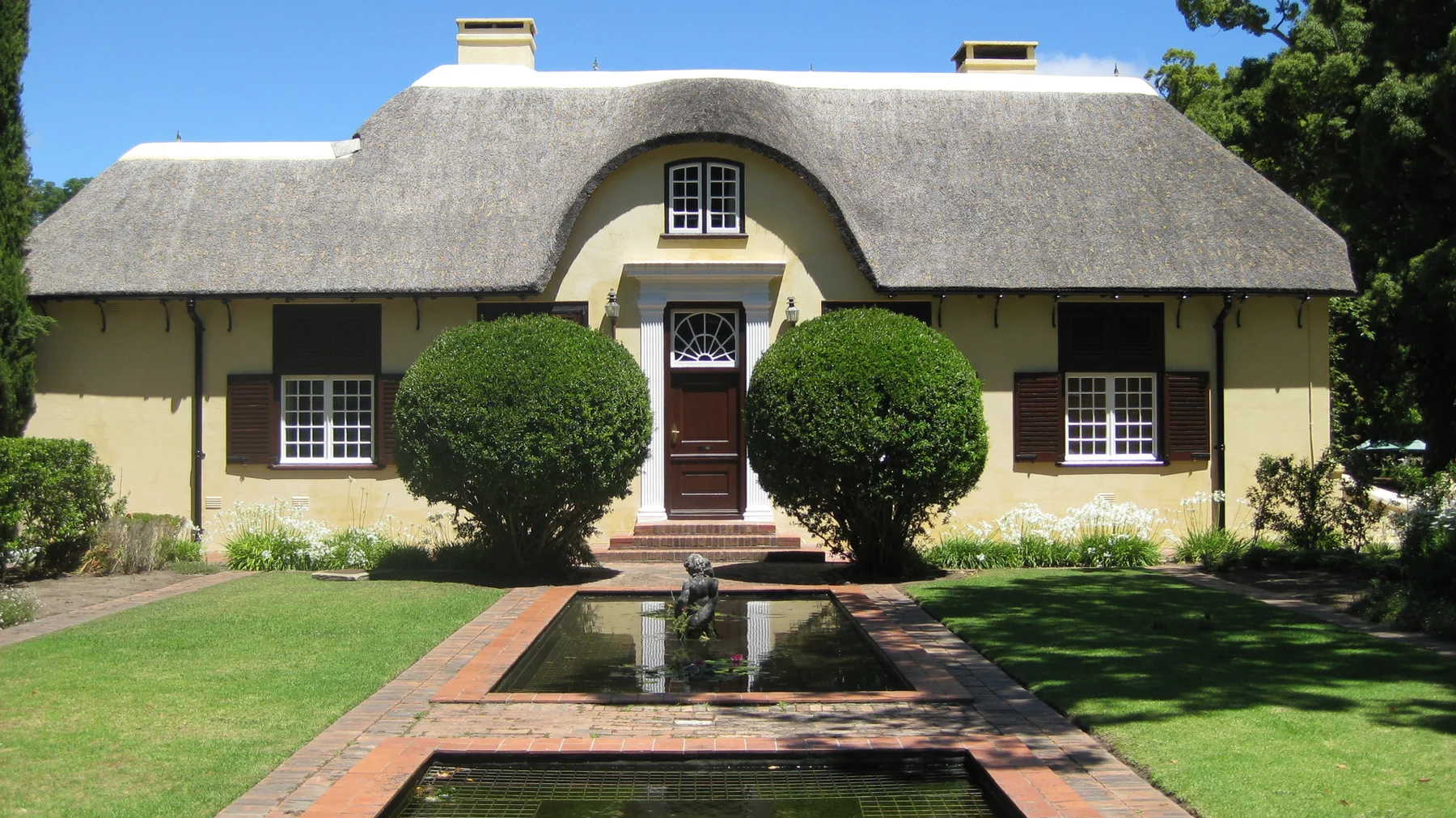 A narrow reflecting pool leads toward a thatched house entrance.