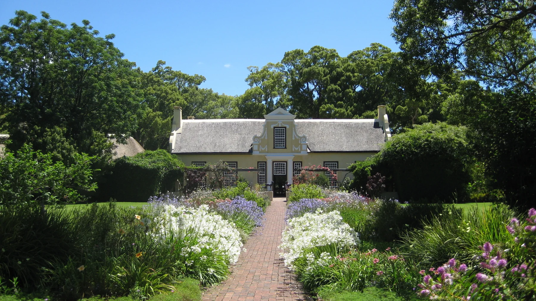 A straight garden path runs between flower borders toward a thatched house.