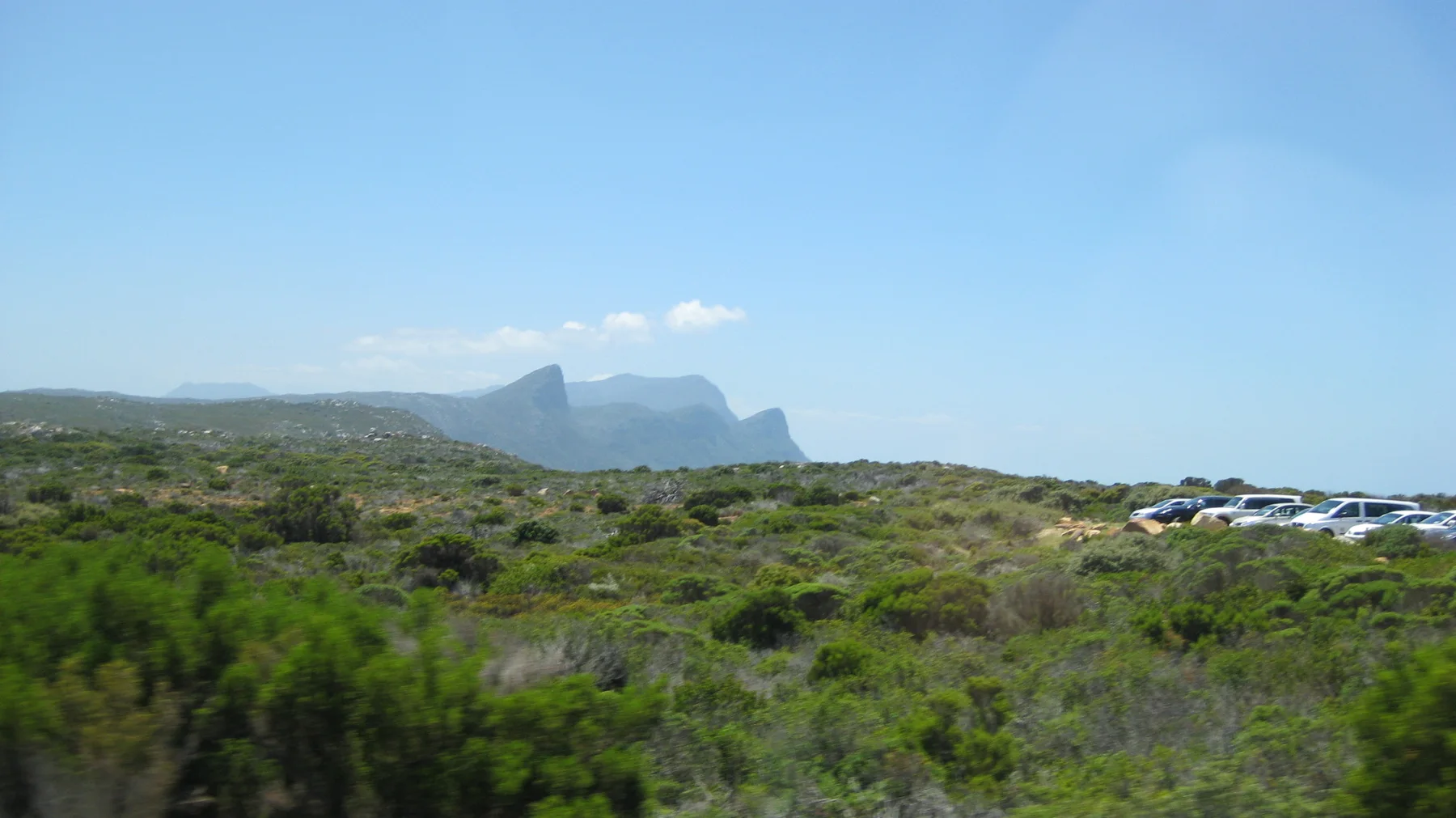 Distant headlands rise beyond open scrub and parked cars.