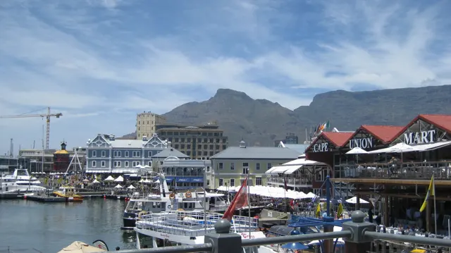 Moored boats sit beneath waterfront buildings and distant mountains.