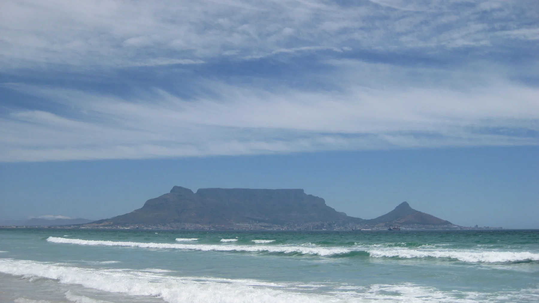 A flat mountain rises beyond surf beneath streaked white clouds.