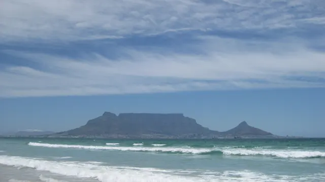 A flat mountain rises beyond surf beneath streaked white clouds.