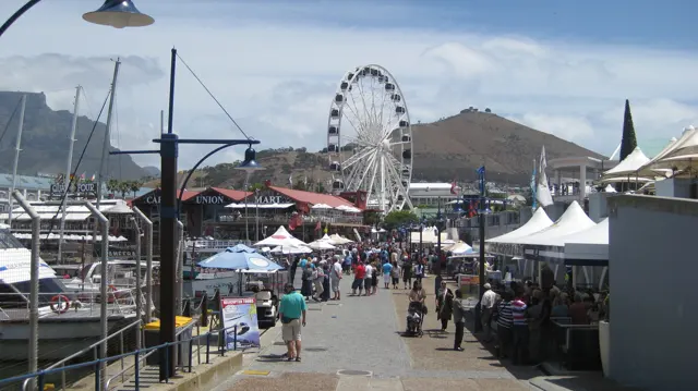 People follow a waterfront walkway toward an observation wheel.