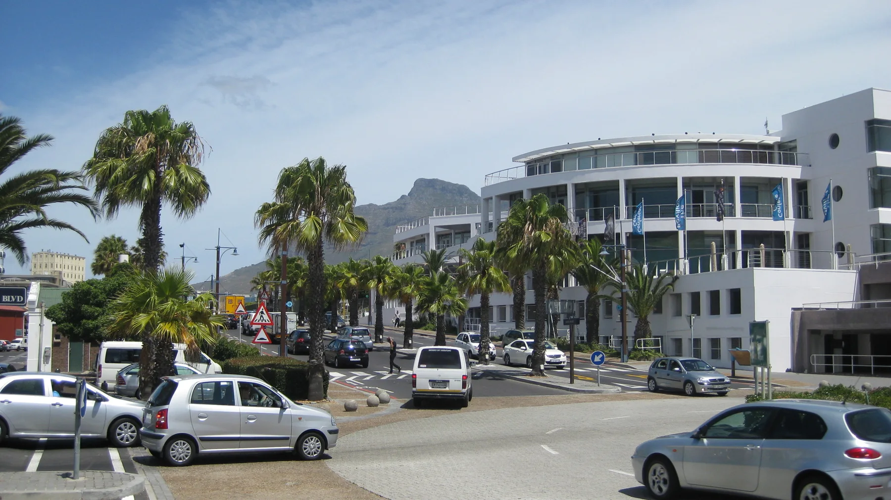 Palms and cars stand before a curved white commercial building.
