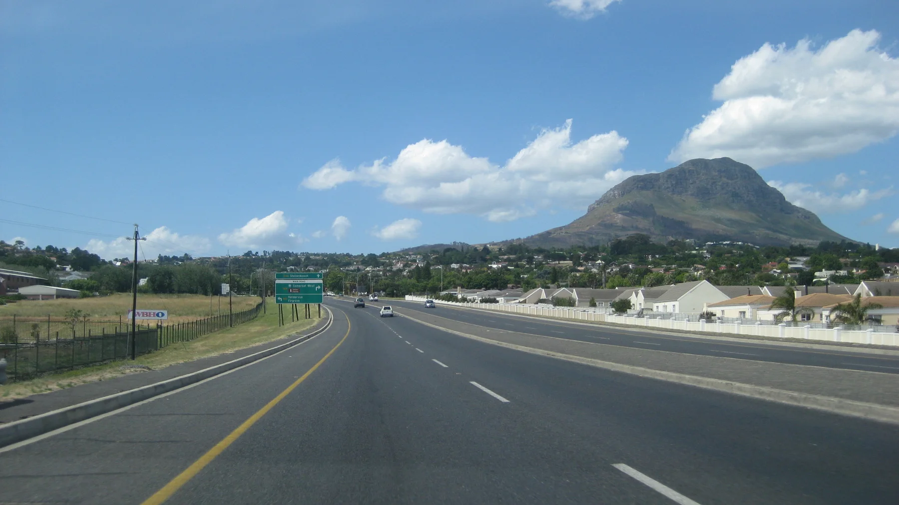 A broad highway curves past open ground below a green hill.