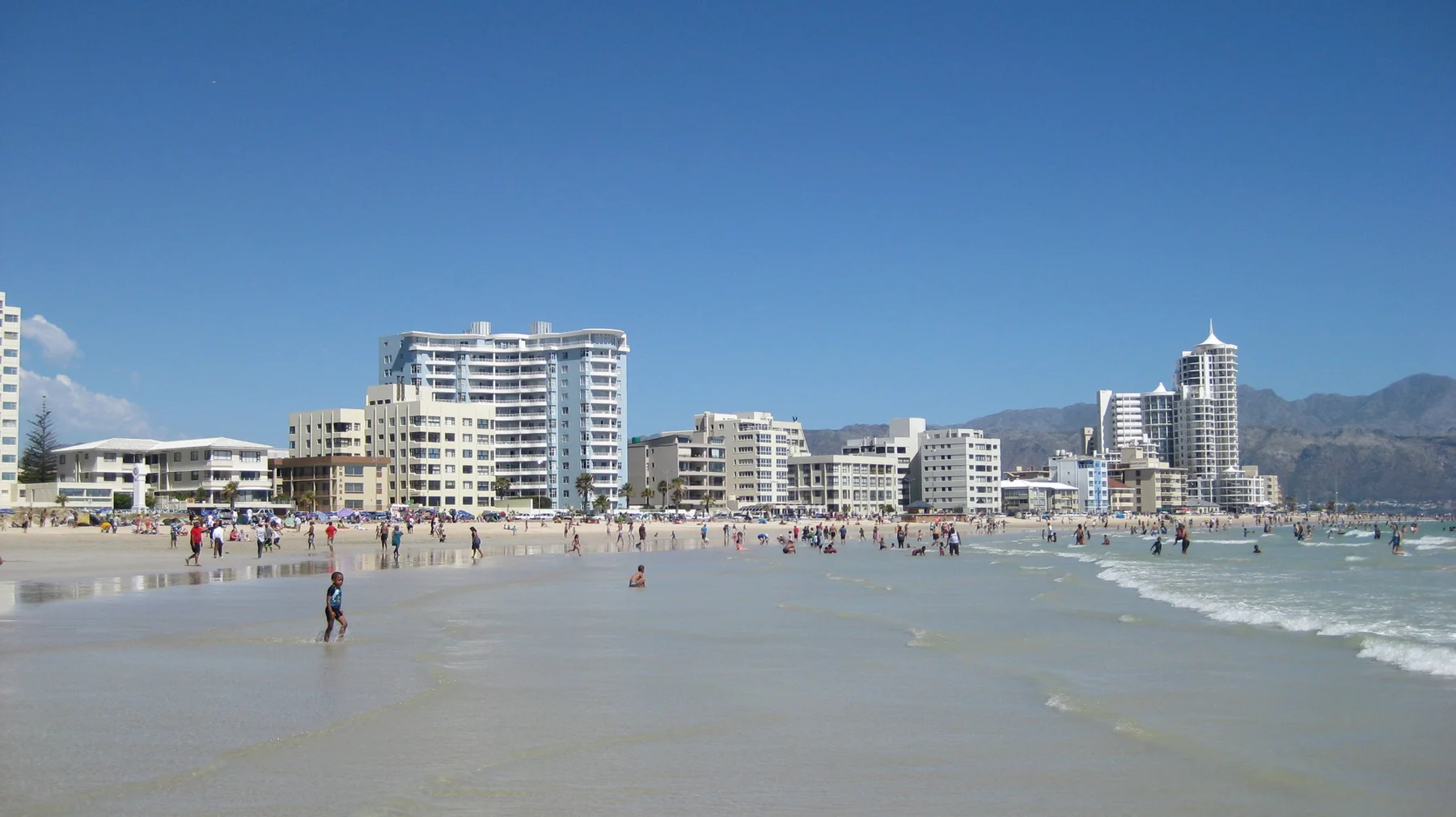 People cross wet reflective sand below waterfront buildings.