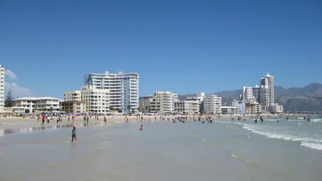 People cross wet reflective sand below waterfront buildings.